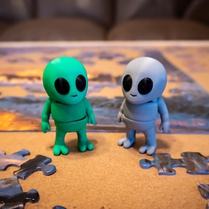 May include: Two small alien figurines, one green and one gray, standing on a wooden surface with puzzle pieces scattered around them.
