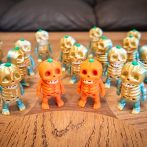 May include: A collection of small standing skeleton figurines with pumpkin-shaped heads. Most figures feature a metallic gold finish on the head and ribcage with teal limbs, while two central figures are solid orange with green stems on top.