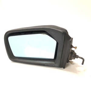Puede incluir: Espejo retrovisor lateral de coche de plástico negro con un espejo de vidrio rectangular. El espejo tiene una palanca de ajuste manual.