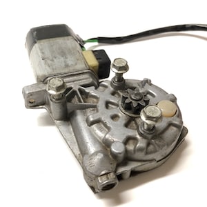 Puede incluir: Un motor de ventana eléctrico plateado y negro con un engranaje y cuatro tornillos. El motor tiene un cable negro conectado.