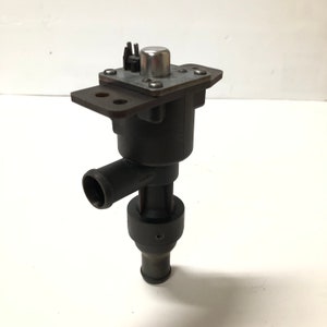 Puede incluir: Una pieza automotriz negra con una placa de metal y una tapa plateada. La pieza tiene un cuerpo cilíndrico con una salida curva y una sección inferior más estrecha. La placa de metal tiene dos orificios de montaje.