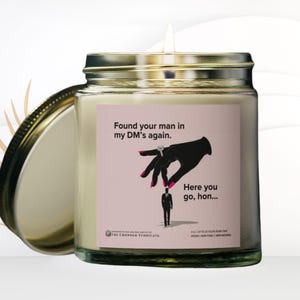 May include: A glass jar candle with a pink label that says "Found your man in my DM's again." and "Here you go, hon..." with an illustration of a hand holding a ring and a small man. The candle is made by The Crowned Syndicate.