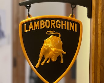 Puede incluir: Un letrero negro y dorado de Lamborghini, con el logotipo de la marca, un toro embistiendo. El letrero, con forma de escudo, tiene un borde dorado y la palabra "Lamborghini" en letras doradas. Cuelga de un soporte de metal negro.
