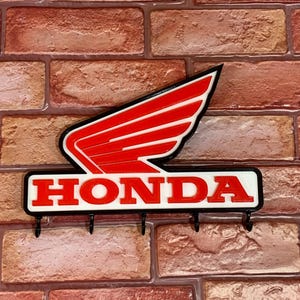 Può includere: Un portachiavi con logo Honda rosso e bianco con cinque ganci. Il logo è un'ala stilizzata con la parola "HONDA" in lettere bianche su sfondo nero.