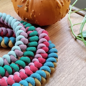 Op de afbeelding: Een close-up van een veelkleurig gevlochten touwspeelgoed. Het touw is gemaakt van vijf verschillende kleuren: mosterdgeel, tealblauw, roze, groen en lichtblauw. Het touw is in een strak patroon gevlochten en is opgerold tot een cirkel.