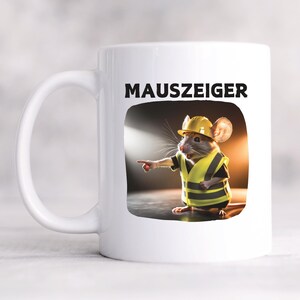 Op de afbeelding: Witte keramische mok met een cartoonmuis met een gele bouwhelm en vest, die met zijn rechterpoot wijst. De mok heeft het woord "MAUSZEIGER" in zwart boven de muisillustratie gedrukt. De mok heeft een wit handvat.