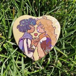 Puede incluir: Un botón en forma de corazón con dos figuras de dibujos animados tomadas de la mano, con un pequeño corazón rojo entre ellas. El botón tiene un fondo amarillo claro con detalles holográficos.