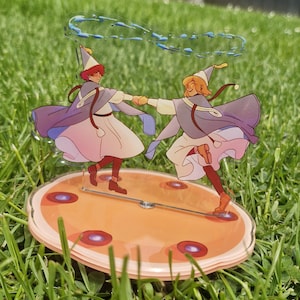 May include: A clear acrylic standee featuring two animated figures in pointed hats, capes, and light-colored robes dancing while holding hands. Above them is a blue swirling magical effect. The figures stand on a round, peach-colored base.