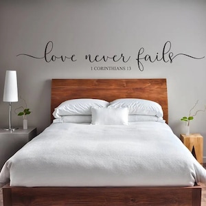 May include: A bedroom with a wooden bed frame and white bedding. The wall above the bed has a black vinyl decal that reads "love never fails 1 Corinthians 13".