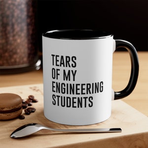 Könnte beinhalten: Weißer Keramikbecher mit schwarzem Innenraum und Henkel. Der Becher trägt den Text "TEARS OF MY ENGINEERING STUDENTS" in fetten schwarzen Buchstaben. Der Becher steht auf einer Holzoberfläche mit einem Löffel, Kaffeebohnen und einem Macaron.