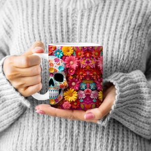 May include: White ceramic mug with a colorful sugar skull and floral design. The skull is decorated with pink, blue, and yellow details. The flowers are pink, red, yellow, and blue.