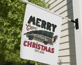 May include: A white Christmas flag with a red and black "Merry Christmas" text and a cartoon image of a station wagon with a Christmas tree on top. The text "Griswold's" is above the station wagon and "EST. 1989" is below the station wagon.