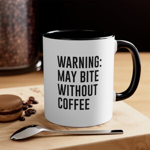 以下が含まれることがあります： 黒色の縁取りと持ち手のある白いコーヒーカップ。カップには「WARNING: MAY BITE WITHOUT COFFEE」という黒い文字が書かれています。