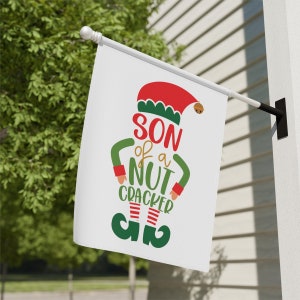 Puede incluir: Una bandera de Navidad blanca con una ilustración de un duende verde y rojo. El duende lleva un gorro rojo con ribete verde y un atuendo a rayas verde y blanco. El texto en la bandera dice "Son of a Nutcracker".