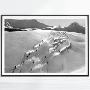 Könnte beinhalten: Ein schwarz-weiß gerahmter Druck, der eine Gruppe von Skifahrern zeigt, die einen schneebedeckten Berghang hinunterfahren. Das Bild zeigt mehrere Personen, die Spuren in den Tiefschnee ziehen und sichtbare Pfade auf dem weißen Untergrund hinterlassen.