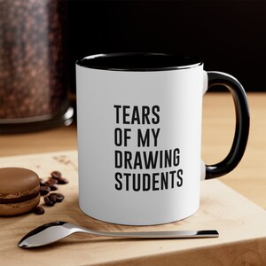 Puede incluir: Taza de café blanca con borde y asa negros. La taza tiene texto negro que dice "Tears of my drawing students".