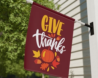 May include: A maroon fabric garden flag with the text "GIVE thanks" in white and yellow lettering. The flag features a stylized pumpkin and autumn leaves in orange, yellow, and brown.