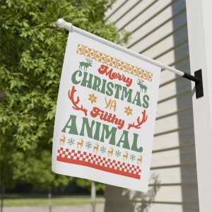May include: A white Christmas flag with the text "Merry Christmas Ya Filthy Animal" in red and green. The flag features reindeer and snowflake designs in orange and white, with a red and white checkered border.