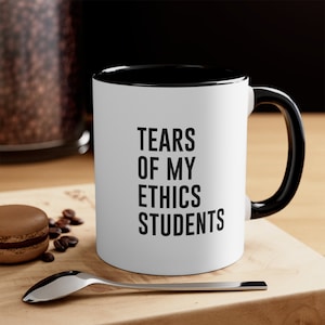 Op de afbeelding: Een witte keramische koffiemok met een zwarte rand en handvat. De mok heeft zwarte tekst met de tekst "Tears of my ethics students".