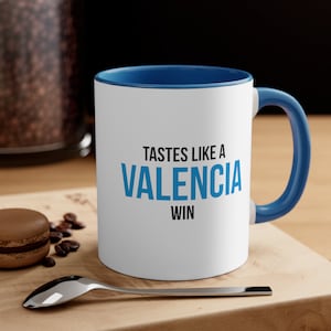 May include: White ceramic mug with a blue rim and handle. The mug has the text "Tastes like a Valencia win" printed on it in black and blue.