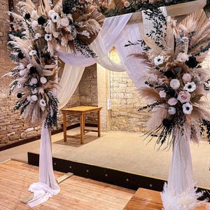 May include: A wedding arch decorated with pampas grass, white fabric, and floral arrangements. The arch is adorned with cream-coloured roses, black flowers, and greenery. The setting is a rustic venue with a wooden floor and stone walls.
