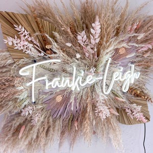 May include: A decorative wall hanging featuring dried pampas grass, feathers, and floral elements in neutral tones. A neon sign spells out "Frankie-Leigh" in a cursive font, adding a modern touch to the natural arrangement.