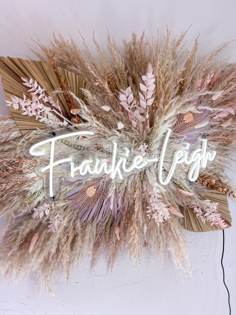 May include: A decorative wall hanging featuring dried pampas grass, feathers, and floral elements in neutral tones. A neon sign spells out "Frankie-Leigh" in a cursive font, adding a modern touch to the natural arrangement.