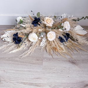 May include: A beige and white pampas grass arrangement with cream and navy blue flowers. The arrangement is on a light wood floor.