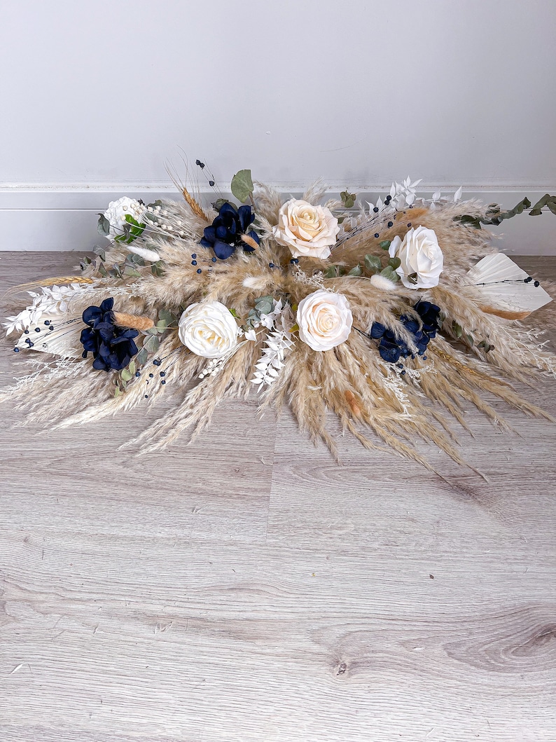 May include: A beige and white pampas grass arrangement with cream and navy blue flowers. The arrangement is on a light wood floor.