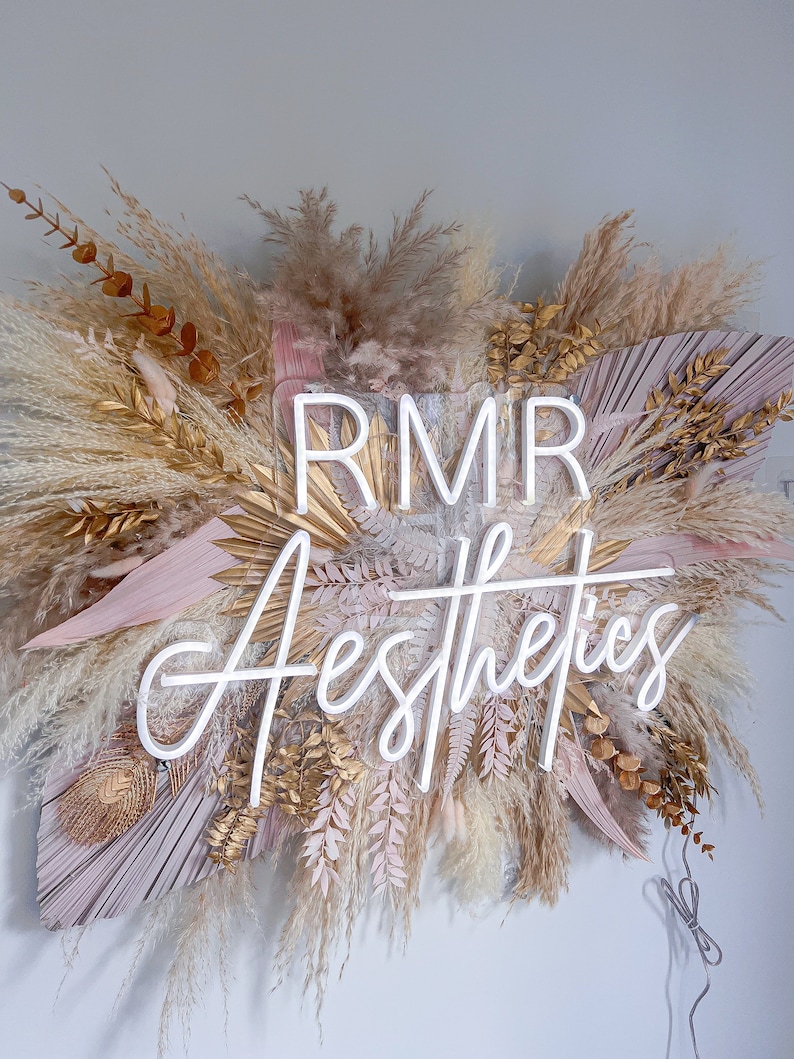 May include: Aesthetic sign featuring the text "RMR Aesthetics" in a white neon light. The sign is surrounded by dried pampas grass, gold-painted leaves, and blush-colored palm leaves, creating a bohemian and natural aesthetic.