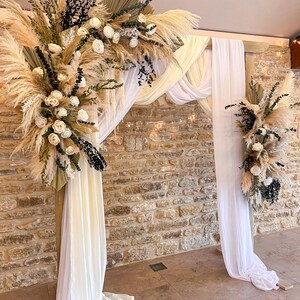 May include: A wooden wedding arch adorned with pampas grass, white roses, and foliage. White fabric drapes from the top and sides of the arch. The backdrop features a stone wall and tiled floor.