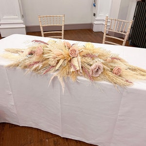 May include: A table is adorned with a floral arrangement featuring pampas grass, roses, and other dried flowers in shades of beige, pink, and cream. Two wooden chairs are positioned behind the table, creating a wedding or event setting. The table is covered with a white tablecloth.
