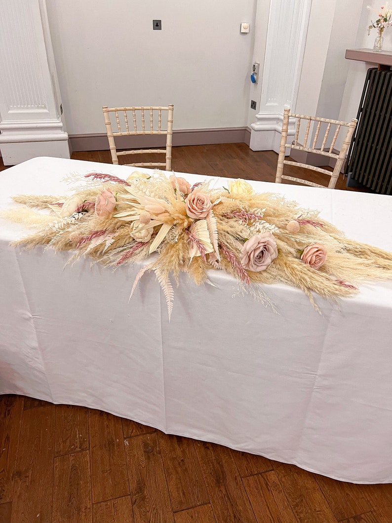 May include: A table is adorned with a floral arrangement featuring pampas grass, roses, and other dried flowers in shades of beige, pink, and cream. Two wooden chairs are positioned behind the table, creating a wedding or event setting. The table is covered with a white tablecloth.