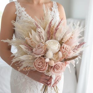 May include: A bridal bouquet featuring a mix of dried and faux flowers in soft pink, cream, and beige tones. The arrangement includes roses, pampas grass, and other textured elements, held together with gold-toned stems.