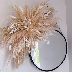 May include: A round mirror with a black frame is hanging on a white wall. A large bouquet of dried pampas grass and white dried flowers is hanging above the mirror.
