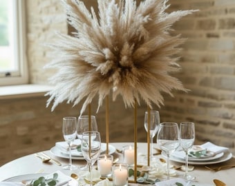 Op de afbeelding: Een prachtig gedekte tafel met een centerpiece van pluizig, beige pampasgras. De tafel is gedekt met witte borden, gouden bestek en wijnglazen. Aangestoken kaarsen en bloemaccenten dragen bij aan de elegante sfeer.