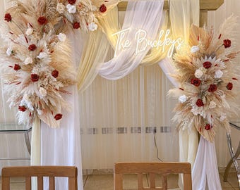 Könnte beinhalten: Hochzeitszeremonie-Dekoration mit einem Holzrahmen, geschmückt mit Pampasgras, roten und weißen Rosen und fließendem weißen Stoff. Das Neonschild zeigt "The Buckleys". Zwei Holzstühle im Vordergrund.