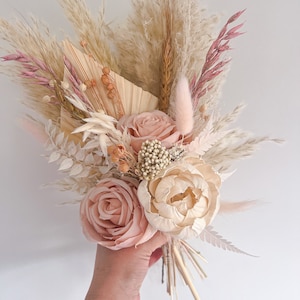 May include: A hand holding a bouquet of dried flowers. The bouquet features large, light pink roses and a cream-colored peony. The arrangement includes pampas grass, wheat stalks, and other dried elements in shades of beige, pink, and cream.