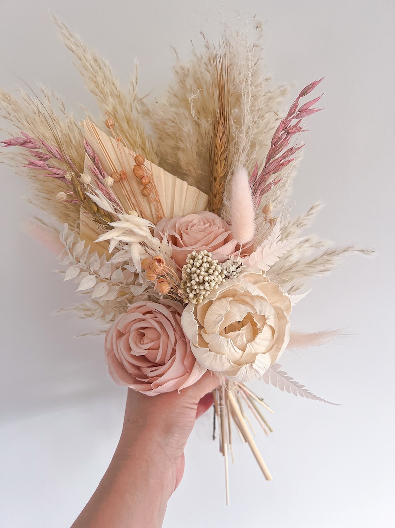 May include: A hand holding a bouquet of dried flowers. The bouquet features large, light pink roses and a cream-colored peony. The arrangement includes pampas grass, wheat stalks, and other dried elements in shades of beige, pink, and cream.