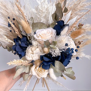 May include: A dried flower bouquet featuring white and cream roses, blue hydrangeas, and dried grasses. The bouquet is tied with natural twine.