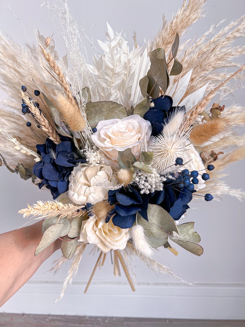 May include: A dried flower bouquet featuring white and cream roses, blue hydrangeas, and dried grasses. The bouquet is tied with natural twine.