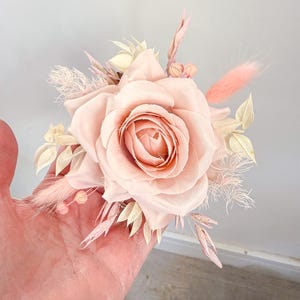 May include: A blush-colored floral arrangement featuring a large rose as the focal point, surrounded by delicate dried flowers and feathery accents. The arrangement is a soft pink hue, perfect for weddings or home decor.