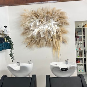 May include: A salon interior featuring two black salon chairs facing white washbasins. Above, a large dried floral arrangement with the word "Serenity" in neon script is mounted on the wall. The room has a clean, modern aesthetic.