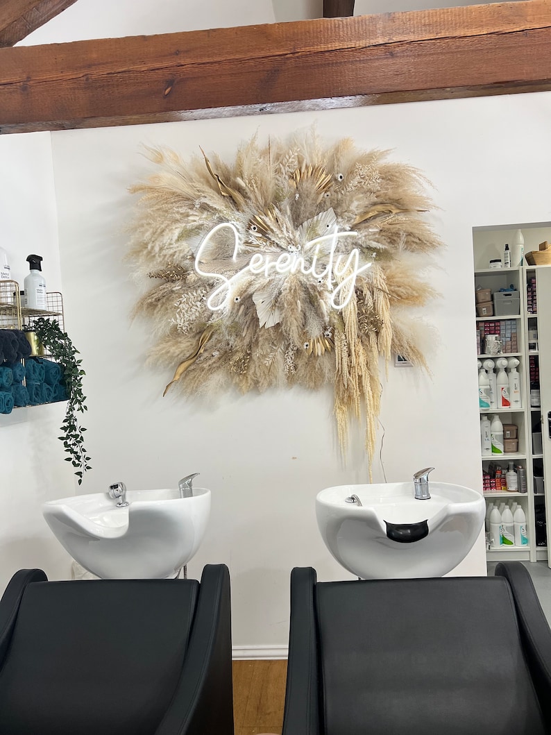 May include: A salon interior featuring two black salon chairs facing white washbasins. Above, a large dried floral arrangement with the word "Serenity" in neon script is mounted on the wall. The room has a clean, modern aesthetic.