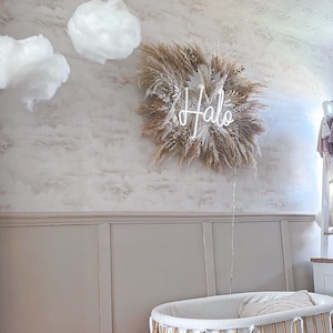 May include: A nursery scene featuring a decorative wall hanging with the word "Halo" in neon, surrounded by dried grasses. The room includes a white bassinet with a bamboo frame and decorative cloud accents.