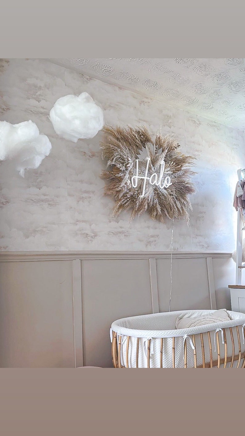 May include: A nursery scene featuring a decorative wall hanging with the word "Halo" in neon, surrounded by dried grasses. The room includes a white bassinet with a bamboo frame and decorative cloud accents.