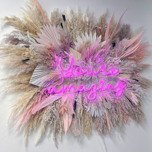 May include: A wall-mounted floral arrangement featuring dried pampas grass, palm leaves, and other decorative elements in shades of pink, beige, and gray. A neon sign in bright pink reads "You're amazing."
