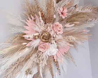 May include: A large wall arrangement of dried and preserved botanicals featuring fluffy pampas grass, pink and white roses, gold-painted palm spears, fern fronds, and small bunny tail grasses. The composition is a mix of cream, beige, and soft pink.