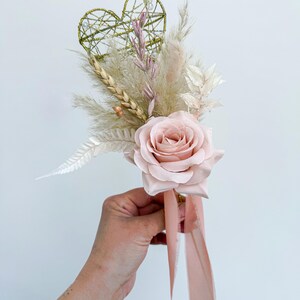 May include: A blush pink rose is the focal point of this floral arrangement. The bouquet includes dried wheat, pampas grass, and a gold wire heart. A blush pink ribbon is tied to the base of the arrangement. The bouquet is held in a person's hand.