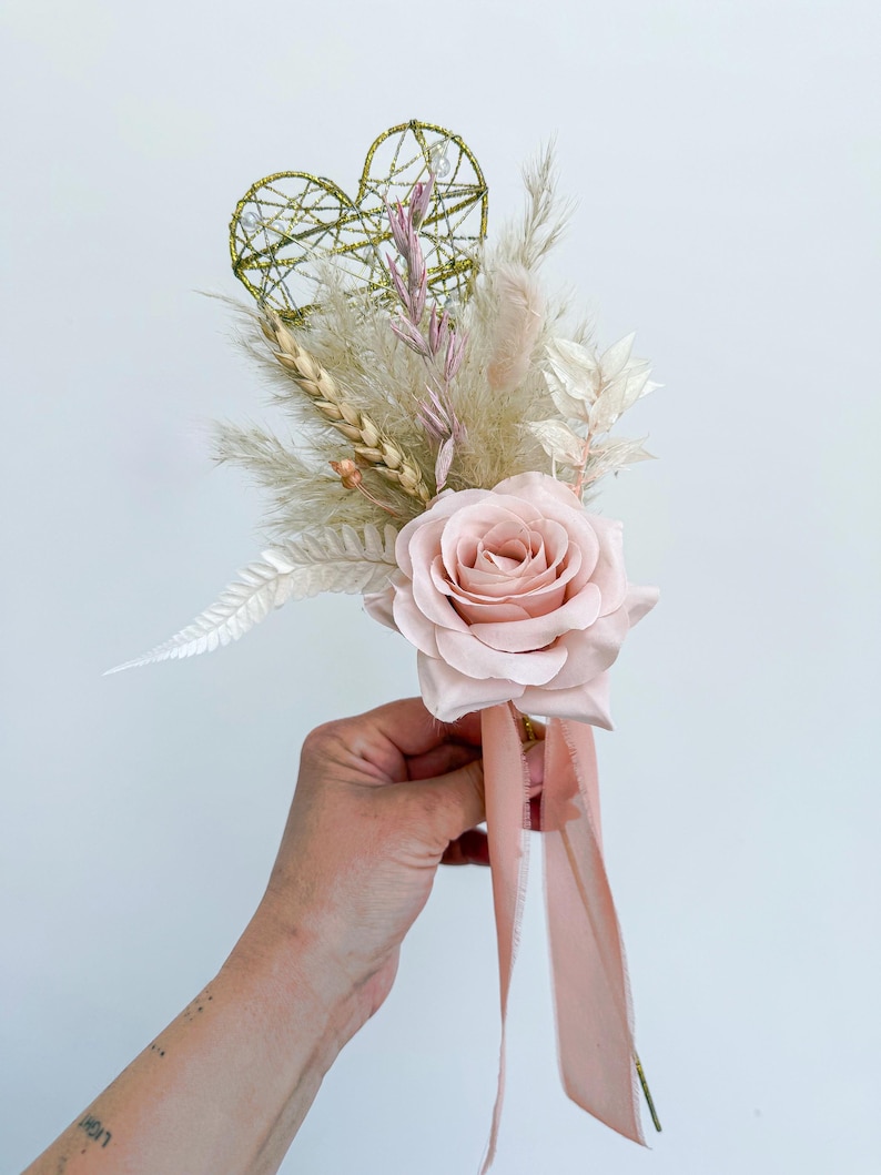 May include: A blush pink rose is the focal point of this floral arrangement. The bouquet includes dried wheat, pampas grass, and a gold wire heart. A blush pink ribbon is tied to the base of the arrangement. The bouquet is held in a person's hand.
